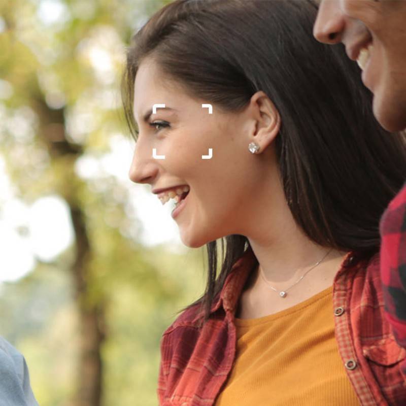 Eine Frau im Freien, die ein rotes Karohemd und ein orangefarbenes Oberteil trägt, lächelt, während sie auf etwas schaut. Der Hintergrund zeigt unscharfe Bäume.