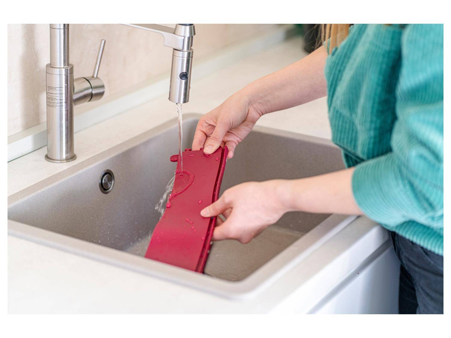Eine Person spült eine rote Silikonmatte unter einem Wasserhahn in einer Spüle, was darauf hindeutet, dass der Gegenstand gereinigt oder gewaschen wird.