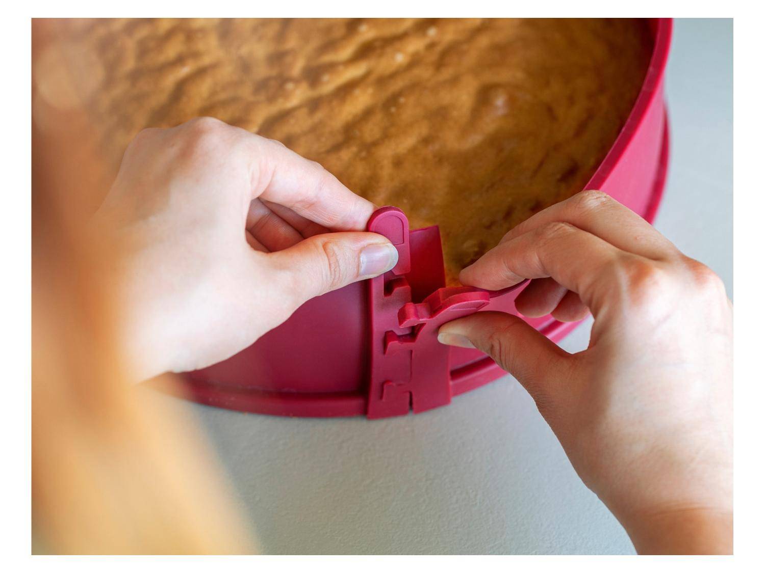 Hände, die eine Springform-Klemme an einer roten Backform lösen, um einen gebackenen Kuchen freizulegen.