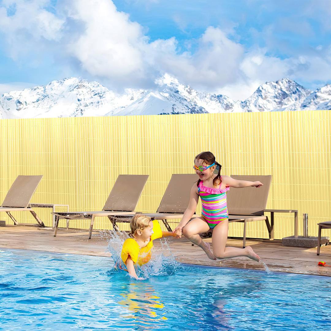 Ein Kind mit gelben Schwimmflügeln schwebt im Wasser und eine Frau in einem Badeanzug genießen einen Swimmingpool mit schneebedeckten Bergen im Hintergrund.