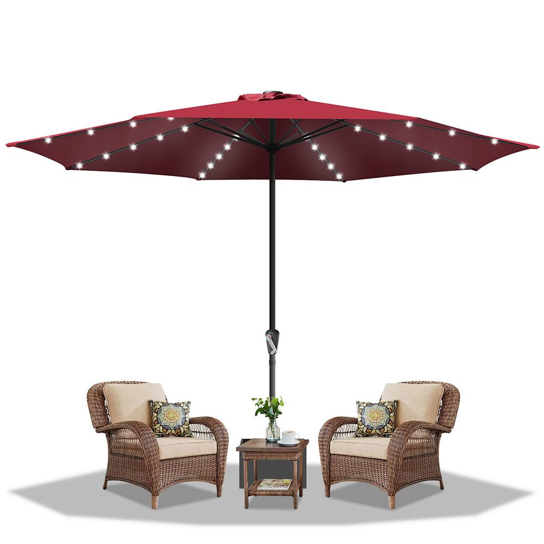 Zwei Korbstühle mit Kissen und dekorativen Kissen stehen sich unter einem großen, beleuchteten burgunderroten Terrassenschirm gegenüber, neben einem kleinen Tisch.