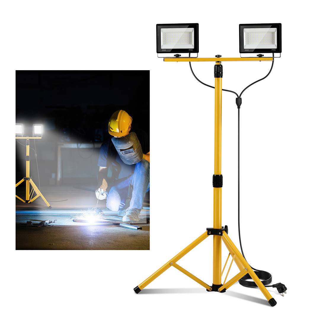 Doppelkopf-Arbeitsleuchte auf einem gelben Dreibeinstativ, mit einer Person in Schutzausrüstung, die in der Dunkelheit schweißt und das Licht zur Sichtbarkeit nutzt.