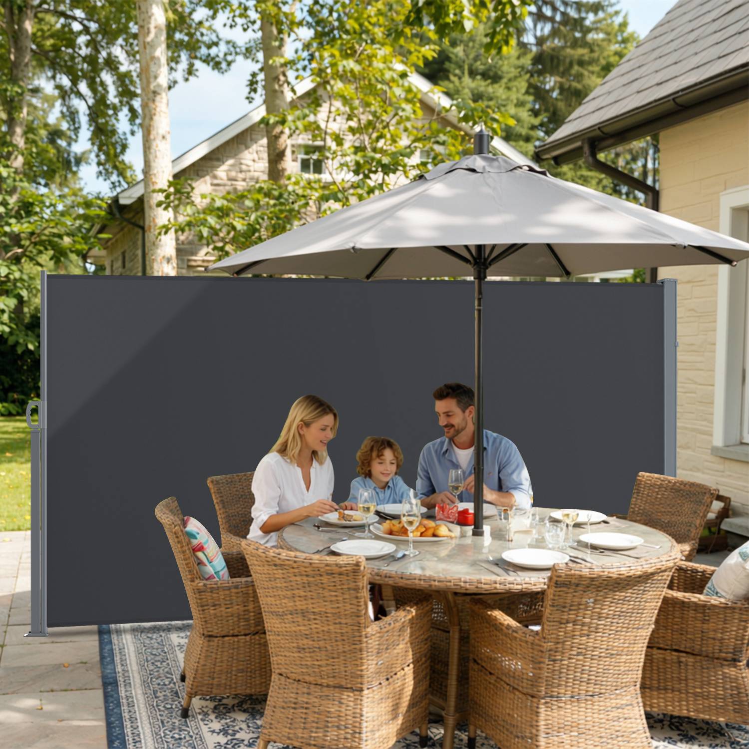 Eine dreiköpfige Familie genießt eine Mahlzeit im Freien an einem runden Tisch mit Korbstühlen unter einem Sonnenschirm, umgeben von Bäumen und einem Haus.
