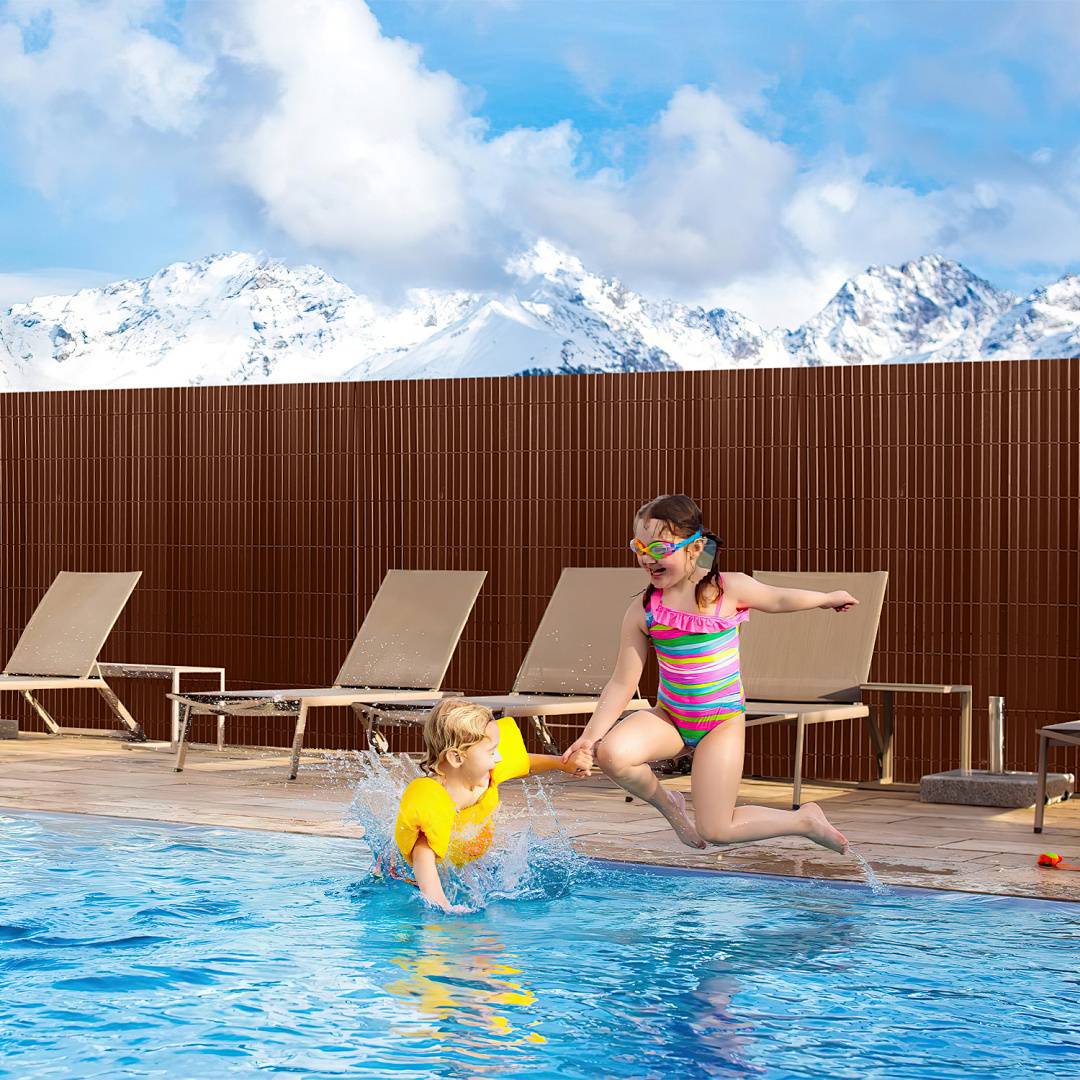Zwei Kinder spielen an einem Pool vor einer Kulisse schneebedeckter Berge. Ein Kind trägt einen Badeanzug und eine Schwimmbrille, das andere hat Schwimmflügel.