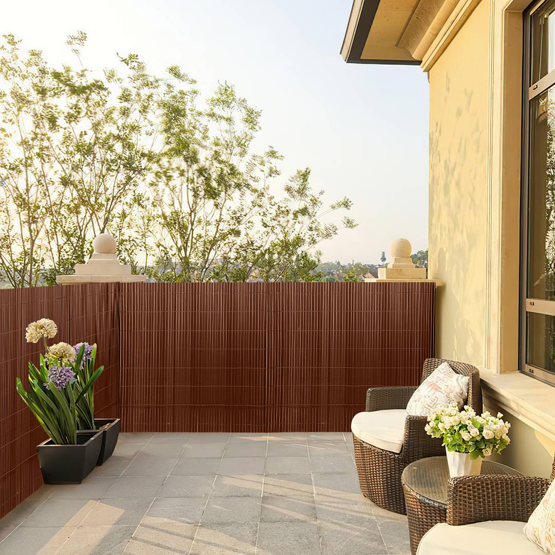 Eine sonnige Terrasse mit Fliesenbelag, Außenmöbeln, Topfpflanzen und Blick auf Bäume hinter einem roten Sichtschutzzaun.