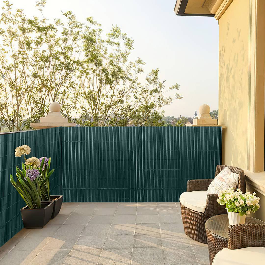 Ein sonniger Balkon mit Korbstühlen, einem kleinen Tisch und Topfpflanzen, umgeben von einem grünen Sichtschutz und mit Blick auf entfernte Bäume.