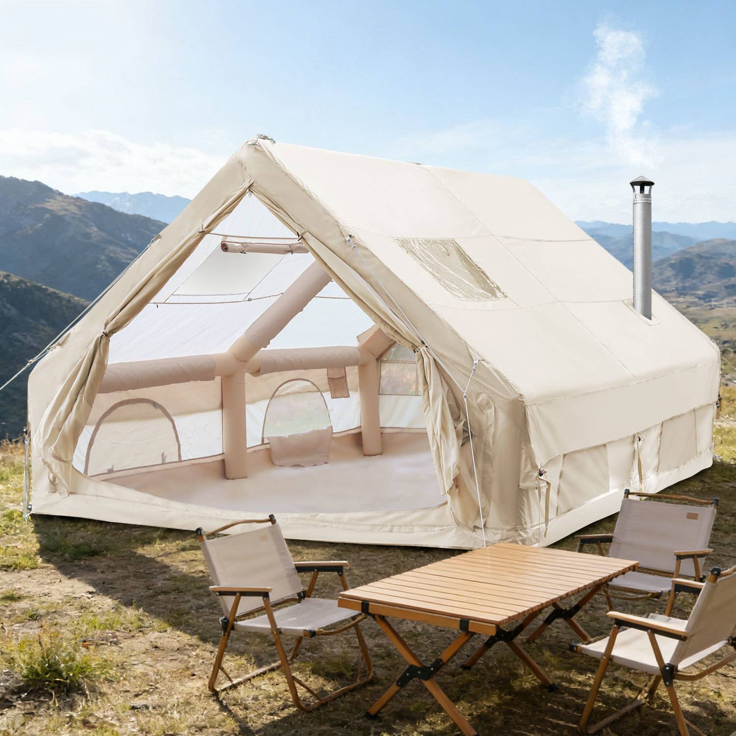 Ein großes cremefarbenes Zelt mit transparenten Abschnitten ist in einer bergigen Gegend aufgestellt. Zwei Campingstühle und ein Holztisch stehen draußen.