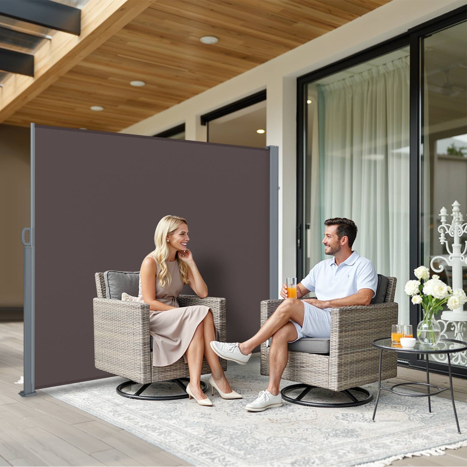 Eine Frau und ein Mann sitzen auf Korbstühlen, getrennt durch einen hohen Raumteiler, auf einer überdachten Terrasse mit hölzerner Decke und Glastüren. Beide lächeln.