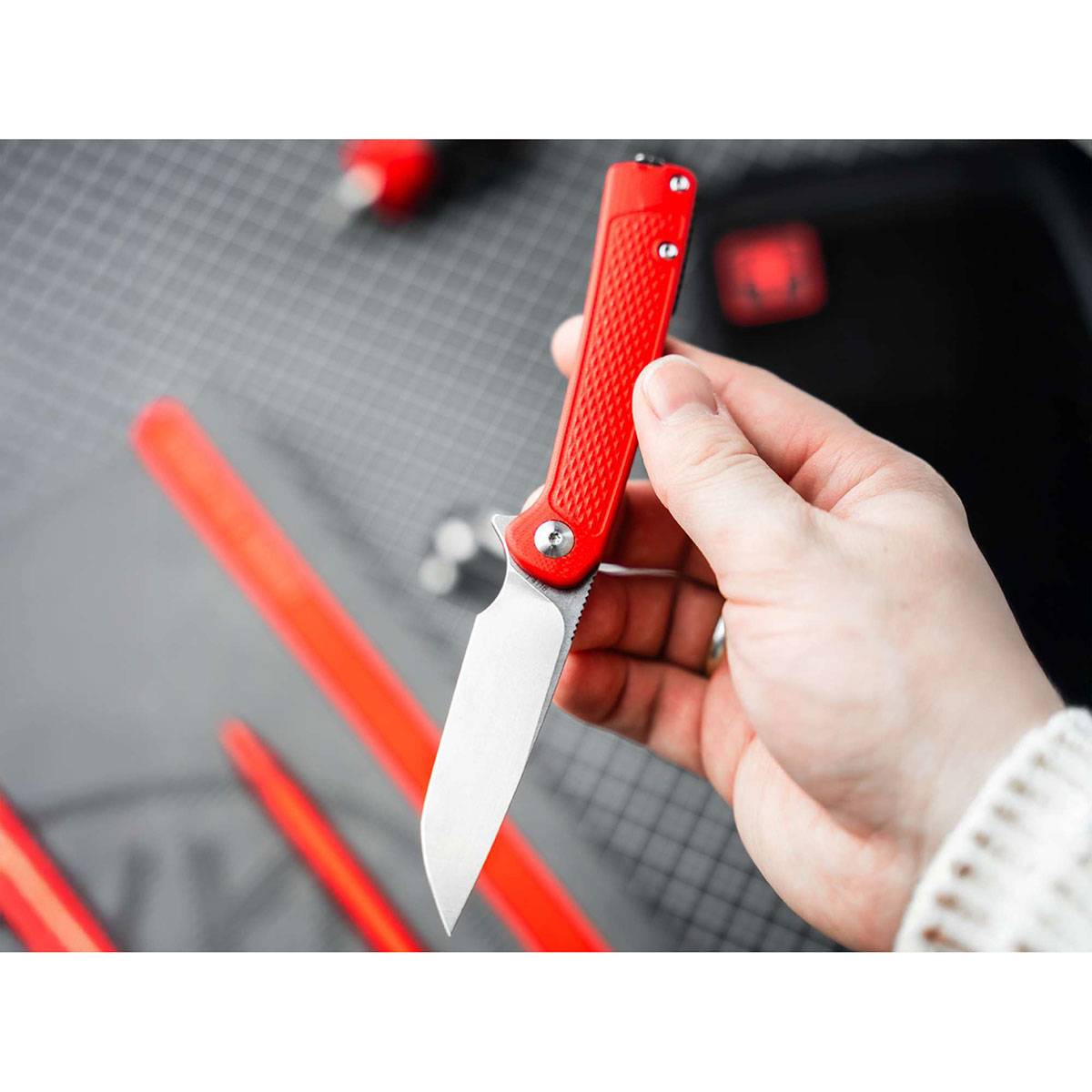 Eine Hand, die ein rotes Universalmesser mit einer silbernen Klinge hält, vor einem unscharfen schwarz-grauem Hintergrund mit Andeutungen von roten Werkzeugen.