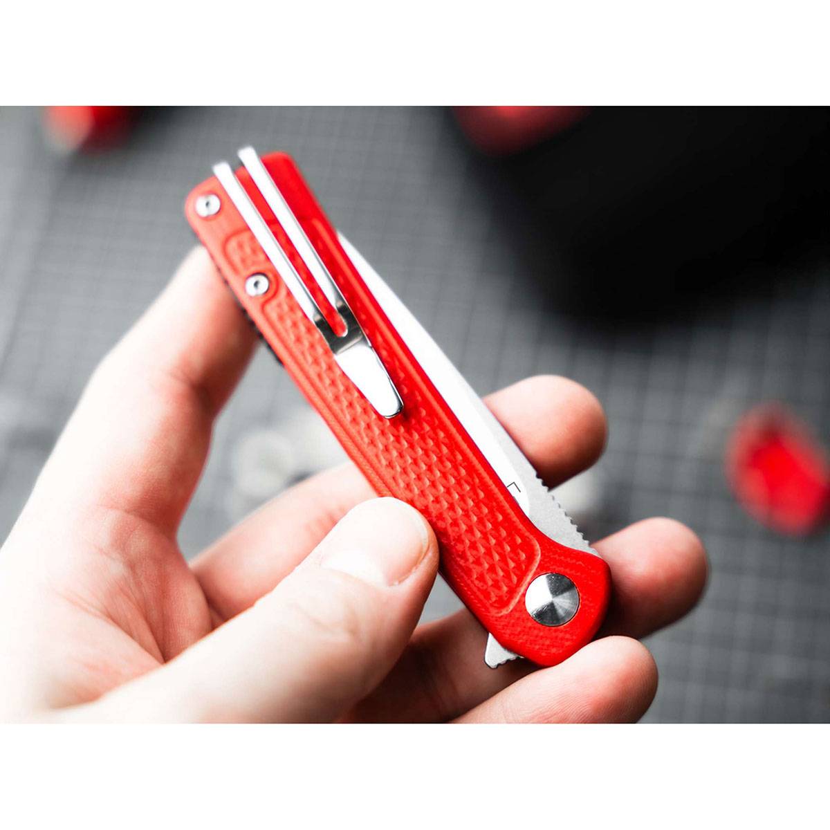 Eine Hand hält ein kleines rotes Multitool mit einer sichtbaren Klinge gegen einen unscharfen Hintergrund mit Andeutungen von roten Objekten.