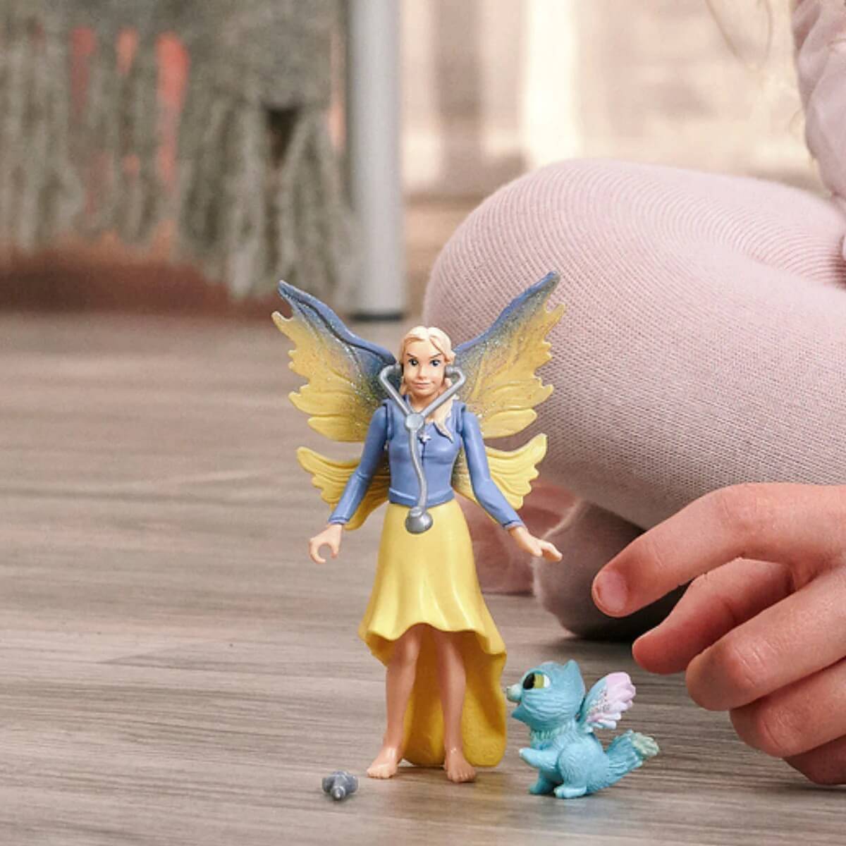 Ein Kind streckt die Hand aus, um eine kleine Feenstatuette und ein blaues Drachenspielzeug auf einem Holzboden zu berühren, was fantasievolles Spiel andeutet.