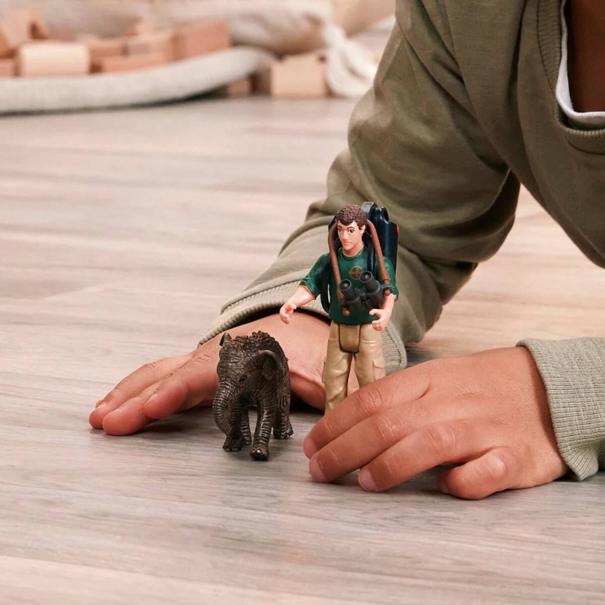 Eine Person auf dem Boden spielt mit kleinen Figuren, darunter ein Elefant und ein Abenteurer mit Rucksack, was eine verspielte, fantasievolle Szene suggeriert.