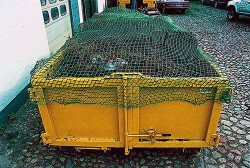 Ein gelber Müllcontainer mit grüner Netzabdeckung ist mit verschiedenen Gegenständen gefüllt und auf einer Kopfsteinpflasteroberfläche neben einem Gebäude platziert.