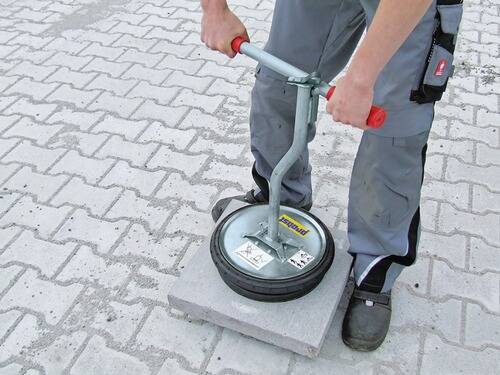 Eine Person demonstriert ein Gerät, das zur Überprüfung der Stabilität von Pflastersteinen verwendet wird. Das Gerät wird auf einem Stein auf einer gepflasterten Oberfläche platziert.