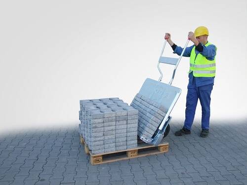 Ein Arbeiter in Schutzausrüstung benutzt eine Handkarre, um eine Palette mit Betonblöcken auf einer gepflasterten Fläche zu heben.