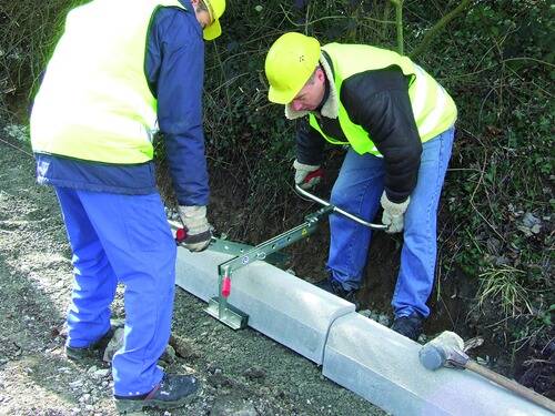 Zwei Arbeiter in gelber Schutzausrüstung verwenden ein Metallwerkzeug, um eine Betonbordsteinkante an der Straßenseite anzuheben und zu positionieren, umgeben von Grünflächen.