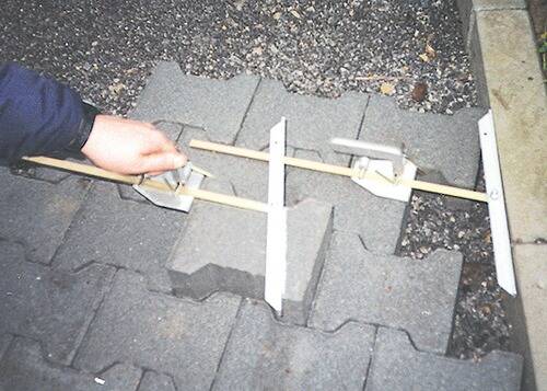 Eine Person, die Abstände zwischen Pflastersteinen mithilfe einer Metallschablone und eines Holzstabes auf einer gepflasterten Oberfläche misst und eine präzise Ausrichtung gewährleistet.