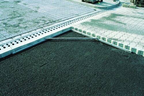 Nahaufnahme eines Betonablaufgitters am Rand eines gepflasterten Parkplatzes, begrenzt durch Verbundsteinplatten und Asphalt.