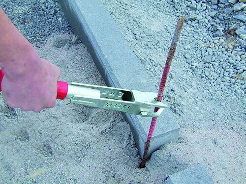 Eine Hand benutzt ein Werkzeug, um eine Metallbewehrung auf einer Baustelle zu biegen, mit Betonblöcken in der Nähe, was einen Bauprozess veranschaulicht.