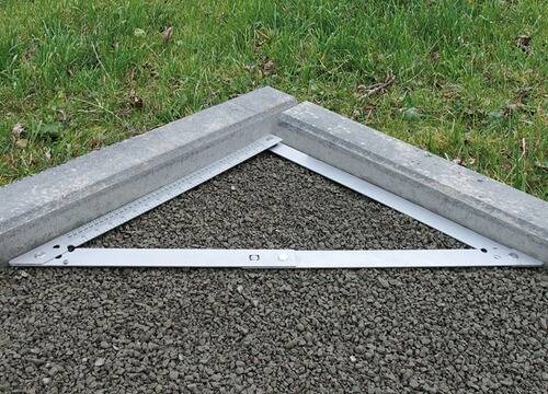 Metalllineal misst rechten Winkel zwischen zwei Betonierten Kanten auf kiesbedecktem Boden.