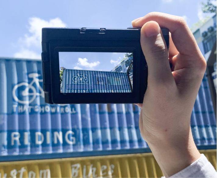 Eine Person hält eine kleine Kamera und rahmt einen blauen Versandcontainer mit einer Fahrradgrafik und sichtbarem Text „Reading
