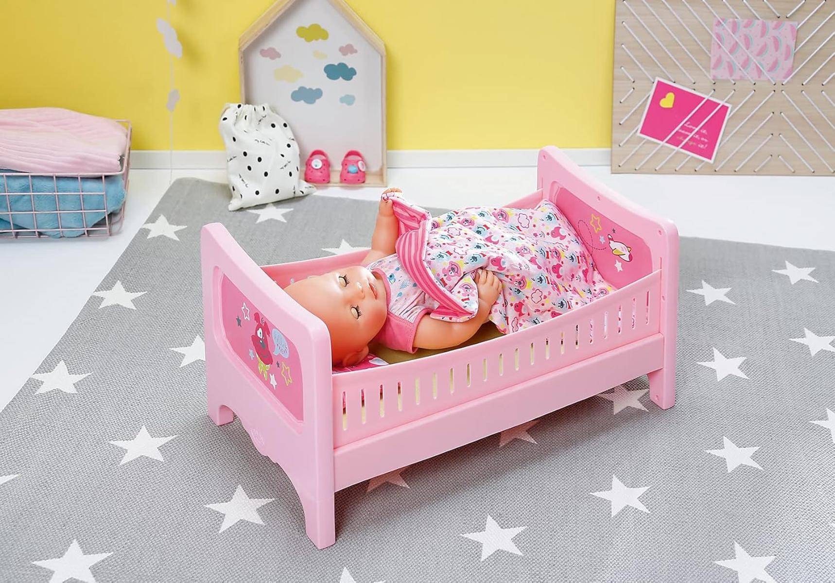 Eine Puppe liegt in einem rosa Spielbettchen mit einer Decke, umgeben von sternförmigen Spielzeugen auf einer grauen Matte in einem farbenfrohen Kinderzimmer.