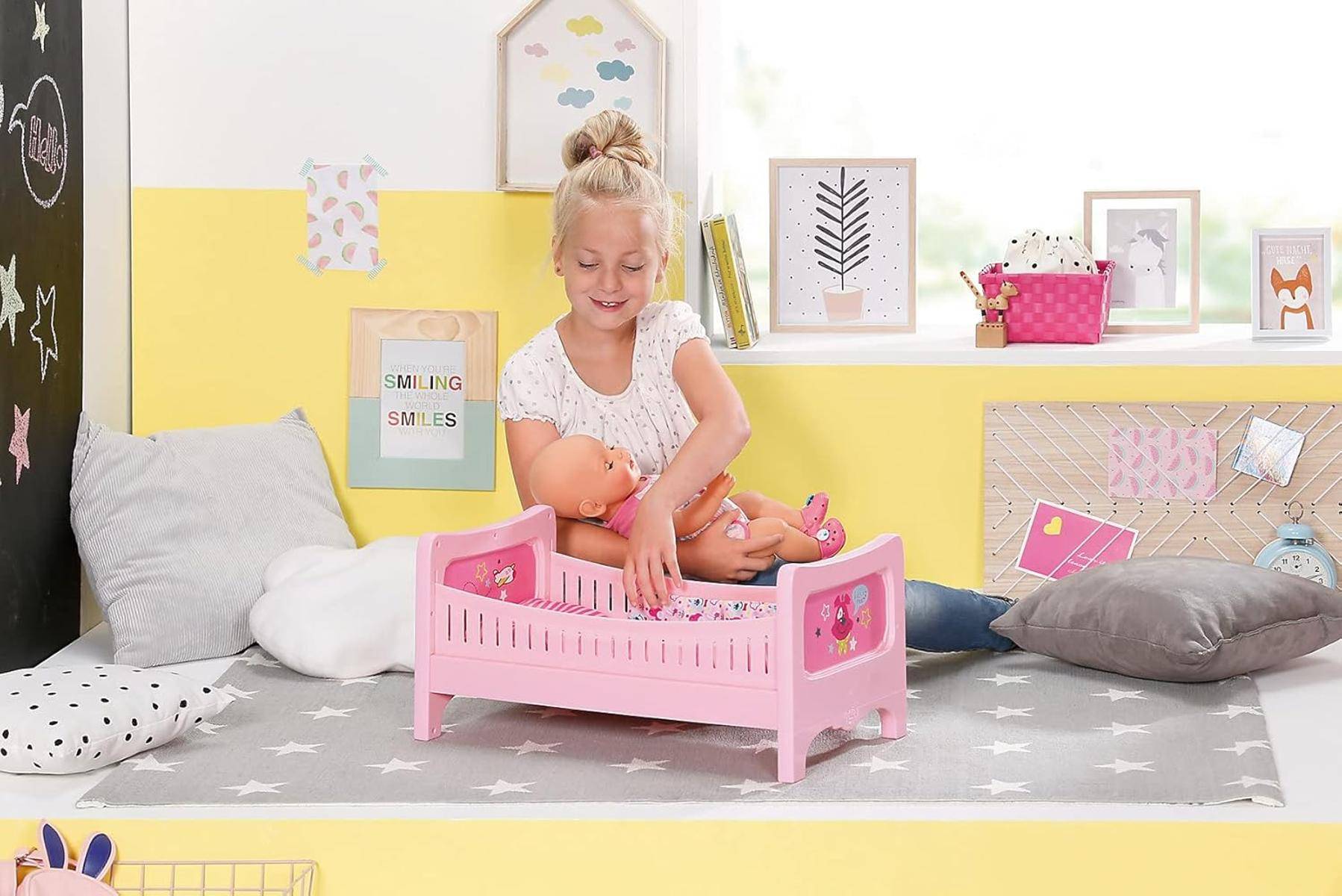 Ein Kind mit blonden Haaren spielt mit einer Babypuppe auf einem rosa Bett in einem Zimmer mit gelben Wänden und dekorativer Kunst an der Wand.