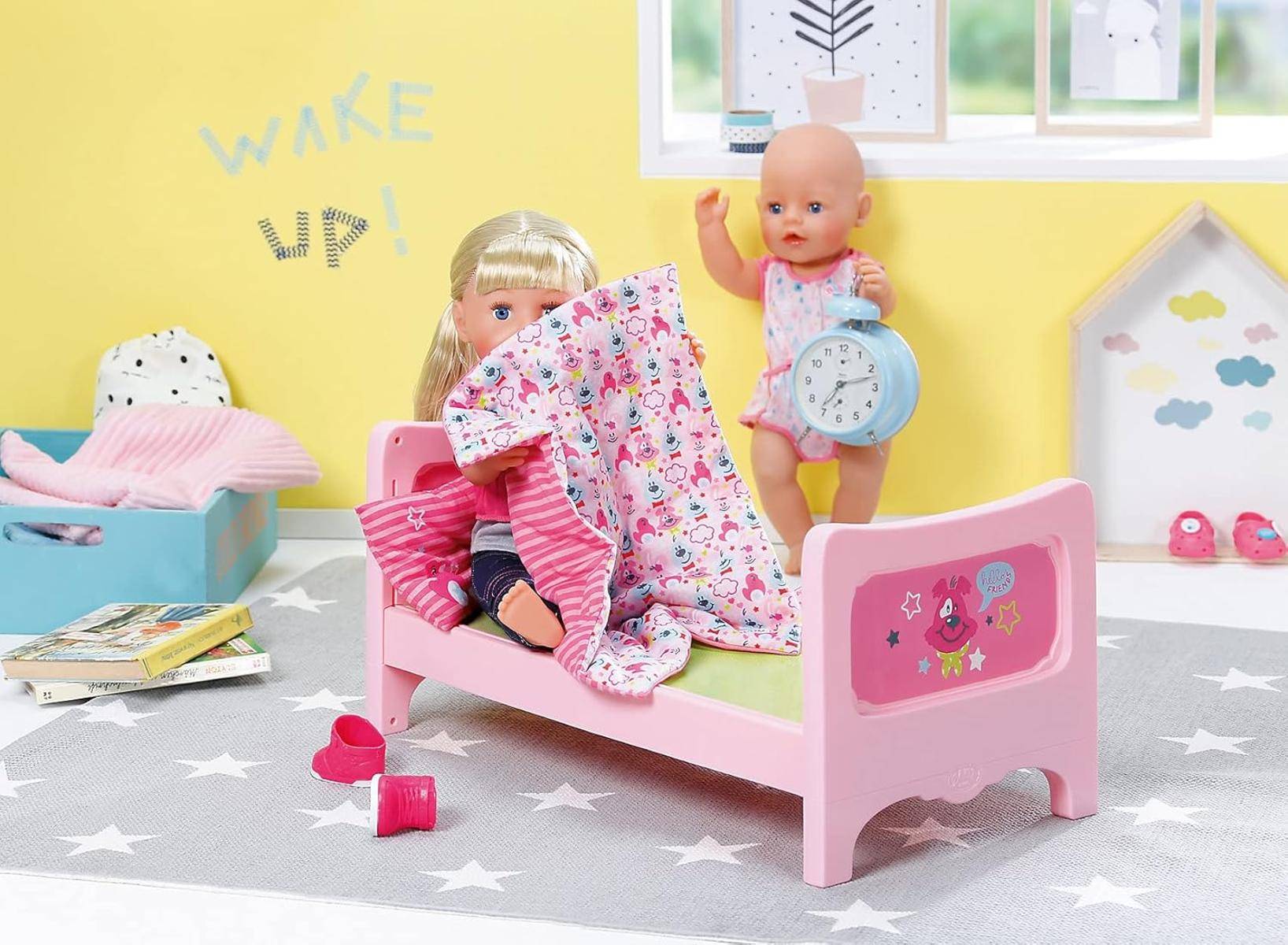 Zwei Puppen in einem Kinderzimmer. Eine liegt auf einem rosa Bett mit einer Decke, und die andere hält eine Uhr. Der Text an der Wand lautet „Wach auf!