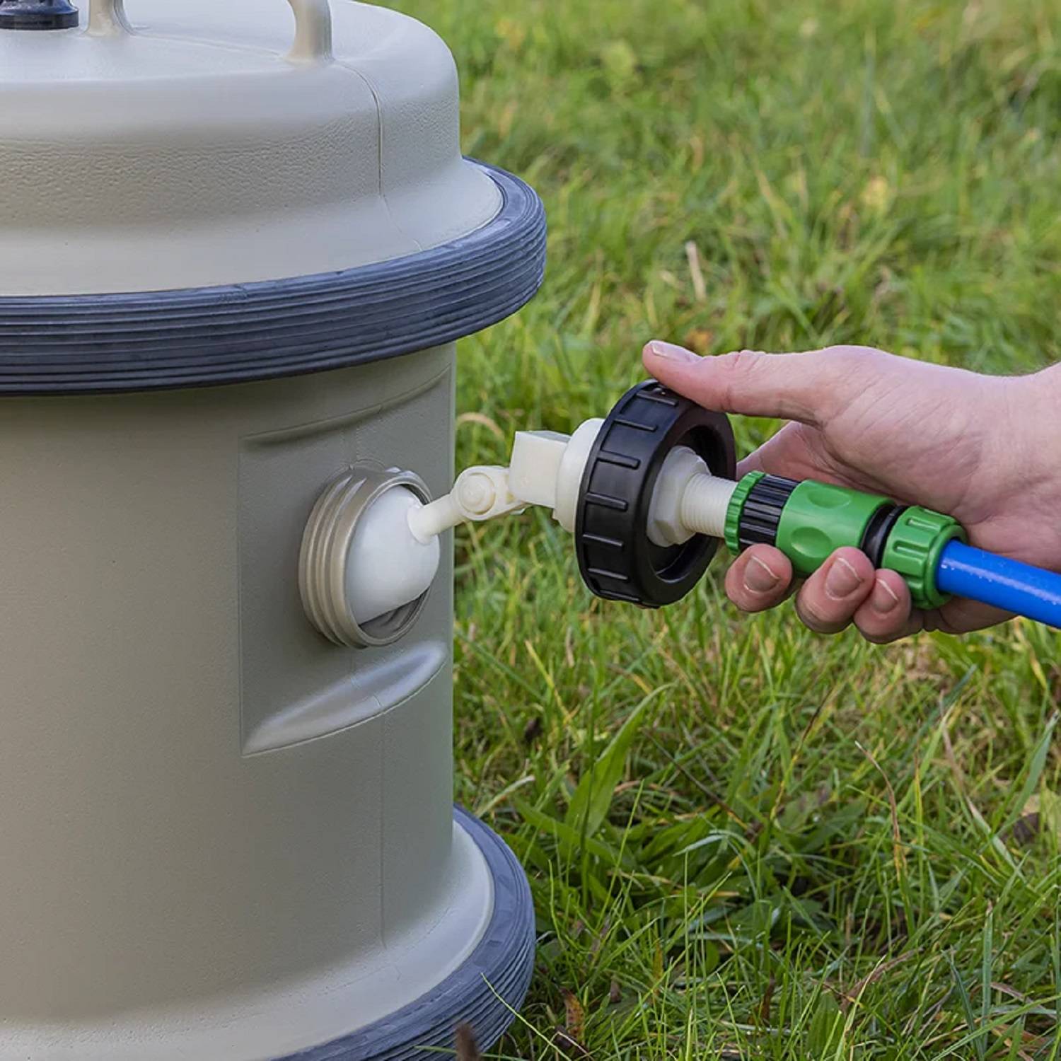 Eine Hand befestigt einen blauen Schlauch an einem Wasserhahn auf einem großen, grauen Behälter, der auf Gras steht, was auf eine Wasserversorgungsanlage im Freien hindeutet.