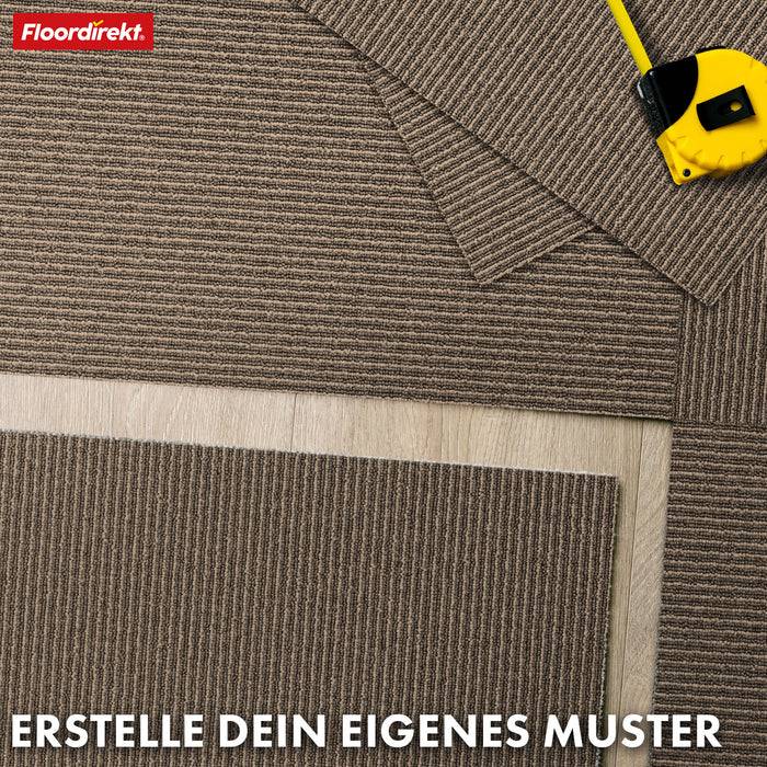 Ein braun und beige gestreifter Teppichmuster ist auf einem Holzboden angeordnet; ein gelbes Messtaband liegt daneben. Der Text lautet „Erstelle dein eigenes Muster.