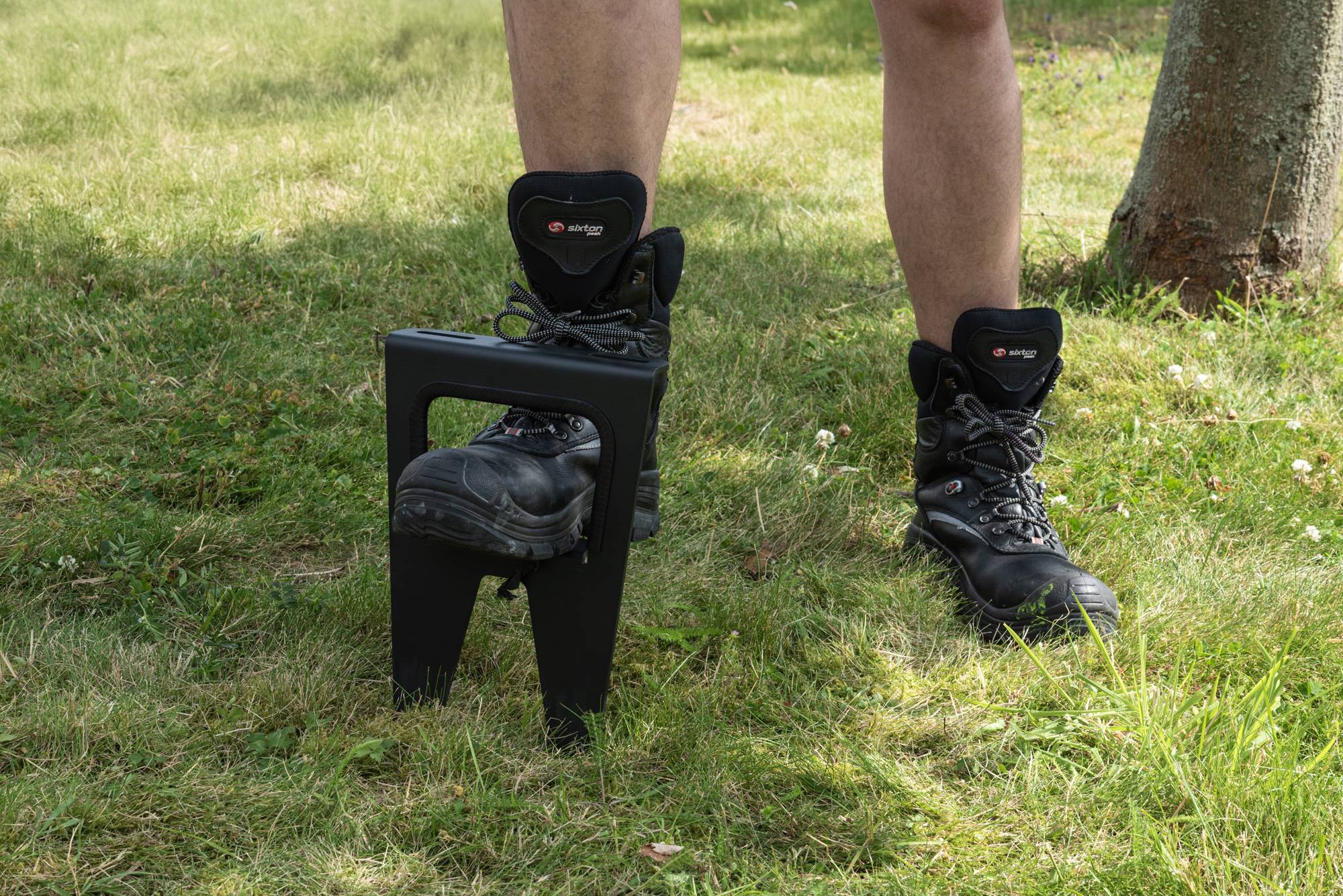 Eine Person, die Stiefel trägt, steht in einem grasbewachsenen Bereich. Ein Stiefel wird von einem schwarzen Metallrahmen gestützt, der für Stabilität oder Bewegungsübungen konzipiert ist.