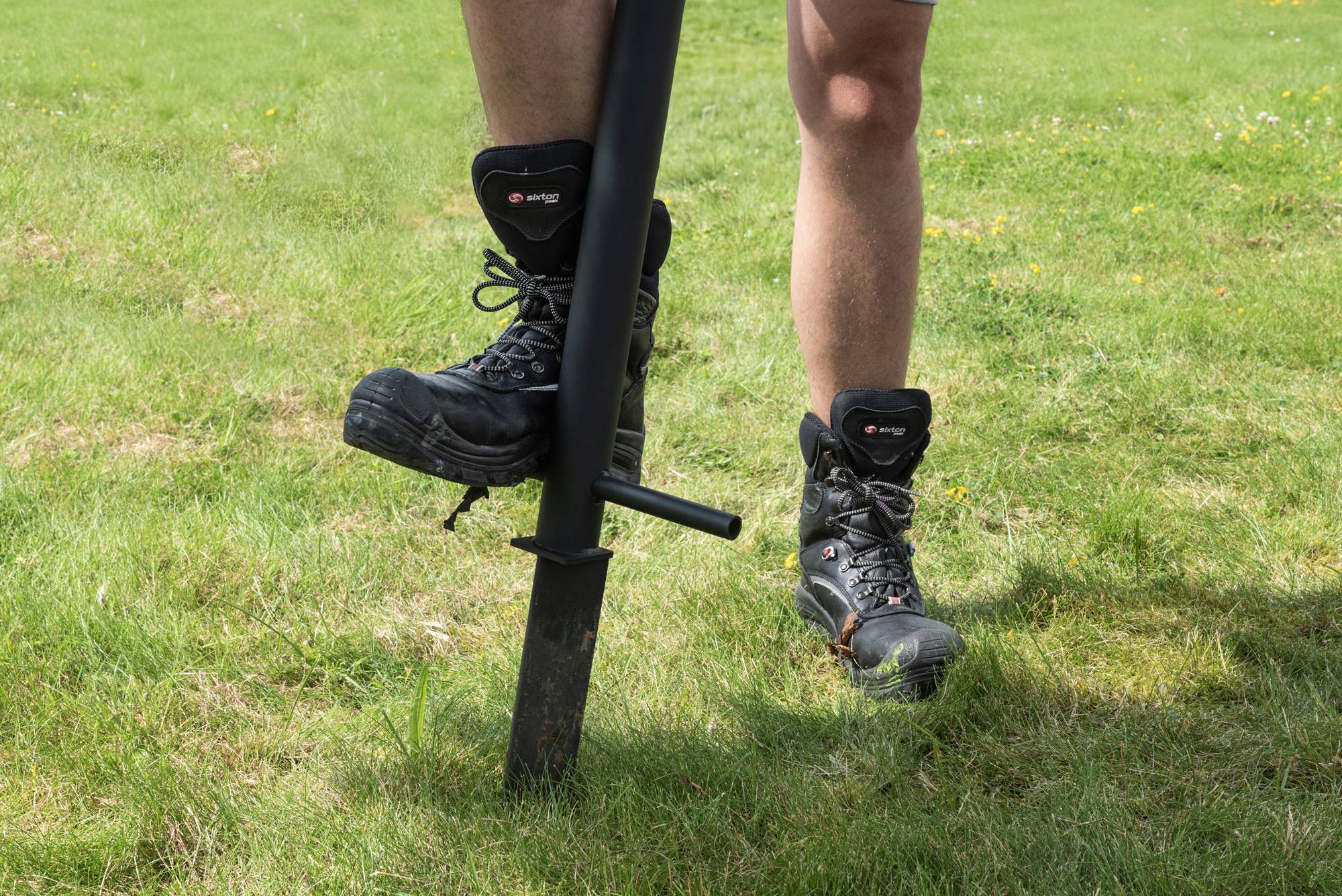 Eine Person, die auf grünem Gras steht, benutzt einen Pfahllochausheber zum Graben, trägt Stiefel und Shorts.