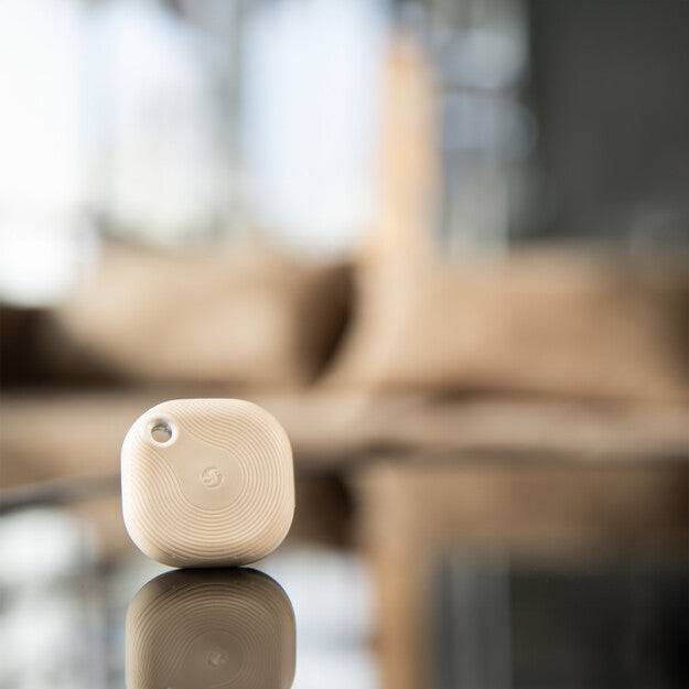 Ein kleines weißes Smart-Home-Gerät mit einem Knopf und einem Spiral-Logo ruht auf einer reflektierenden Oberfläche vor einem unscharfen Hintergrund aus Kissen.