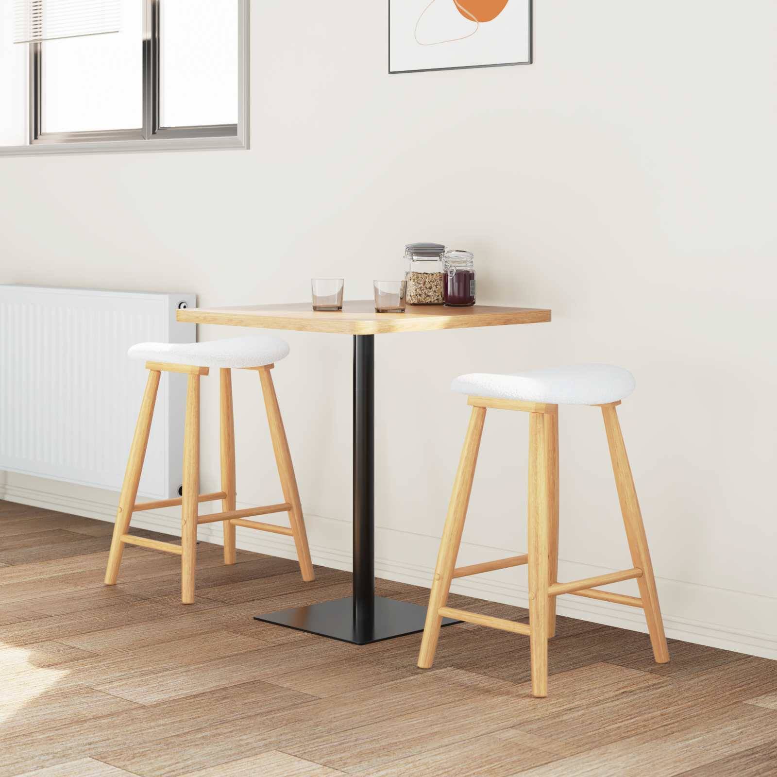 Ein minimalistischer Essbereich mit einem kleinen Holztisch, zwei Hockern, einem Fenster und gerahmten Kunstwerken an der Wand. Natürliches Licht erfüllt den Raum.