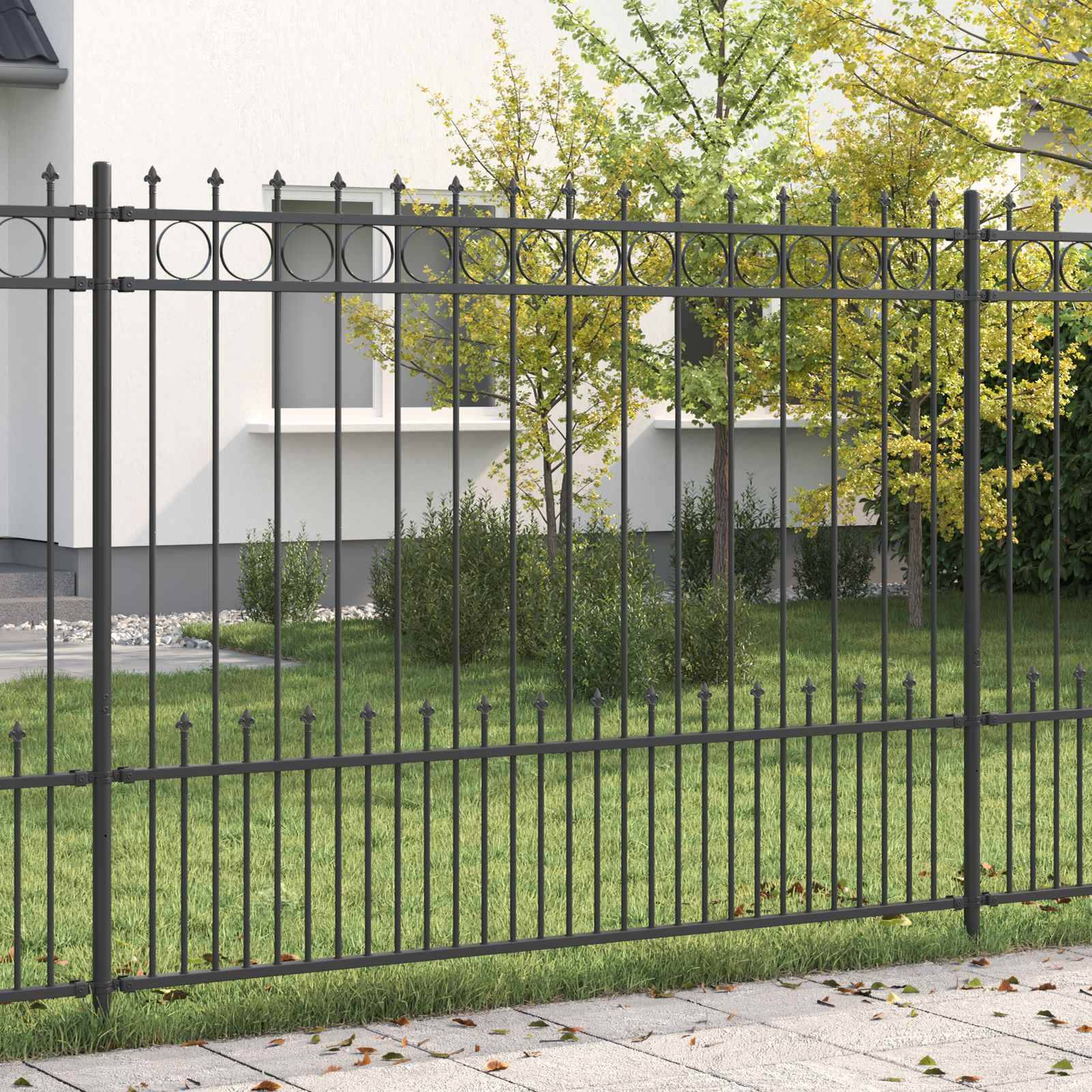 Ein Schmiedeeisenzaun mit dekorativen Elementen säumt einen grasbewachsenen Hof und einen gepflasterten Weg, mit einem Haus und Bäumen im Hintergrund.