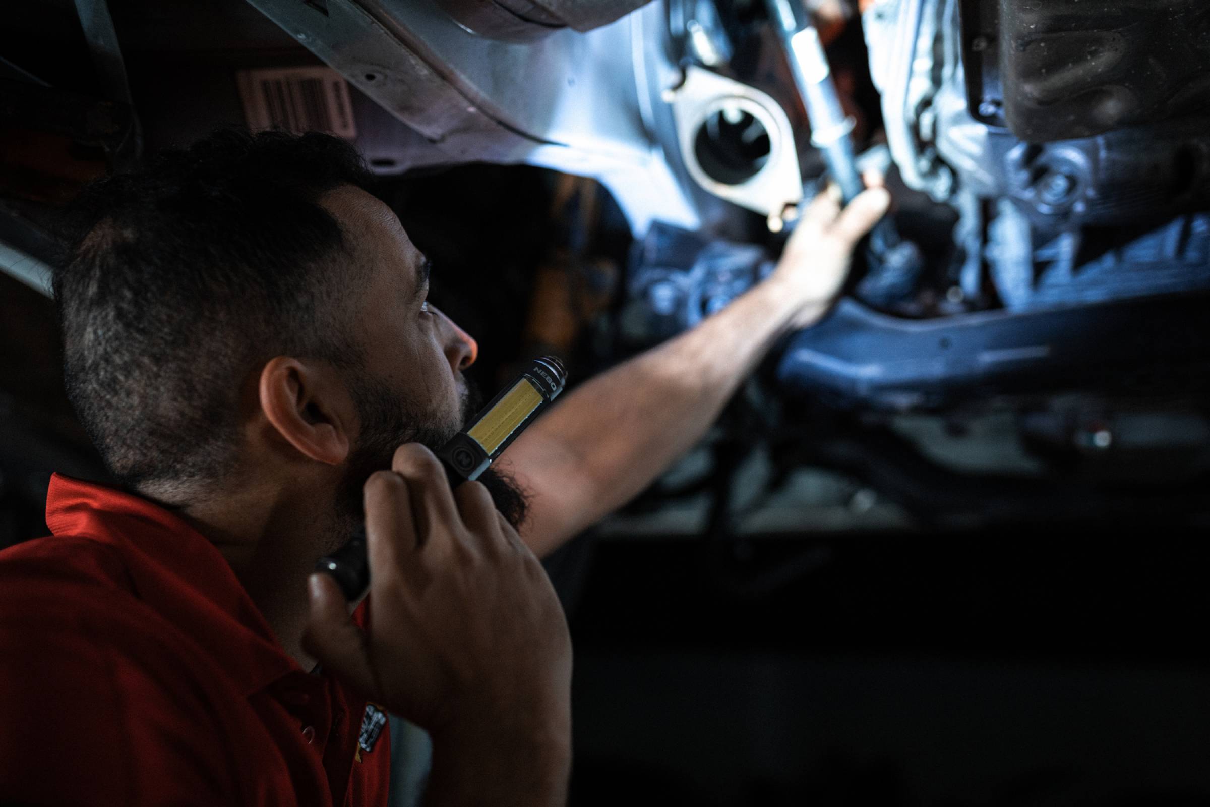 Mechaniker inspiziert einen Automotor mit einer Taschenlampe in einer dunklen Garage und greift unter die Motorhaube.