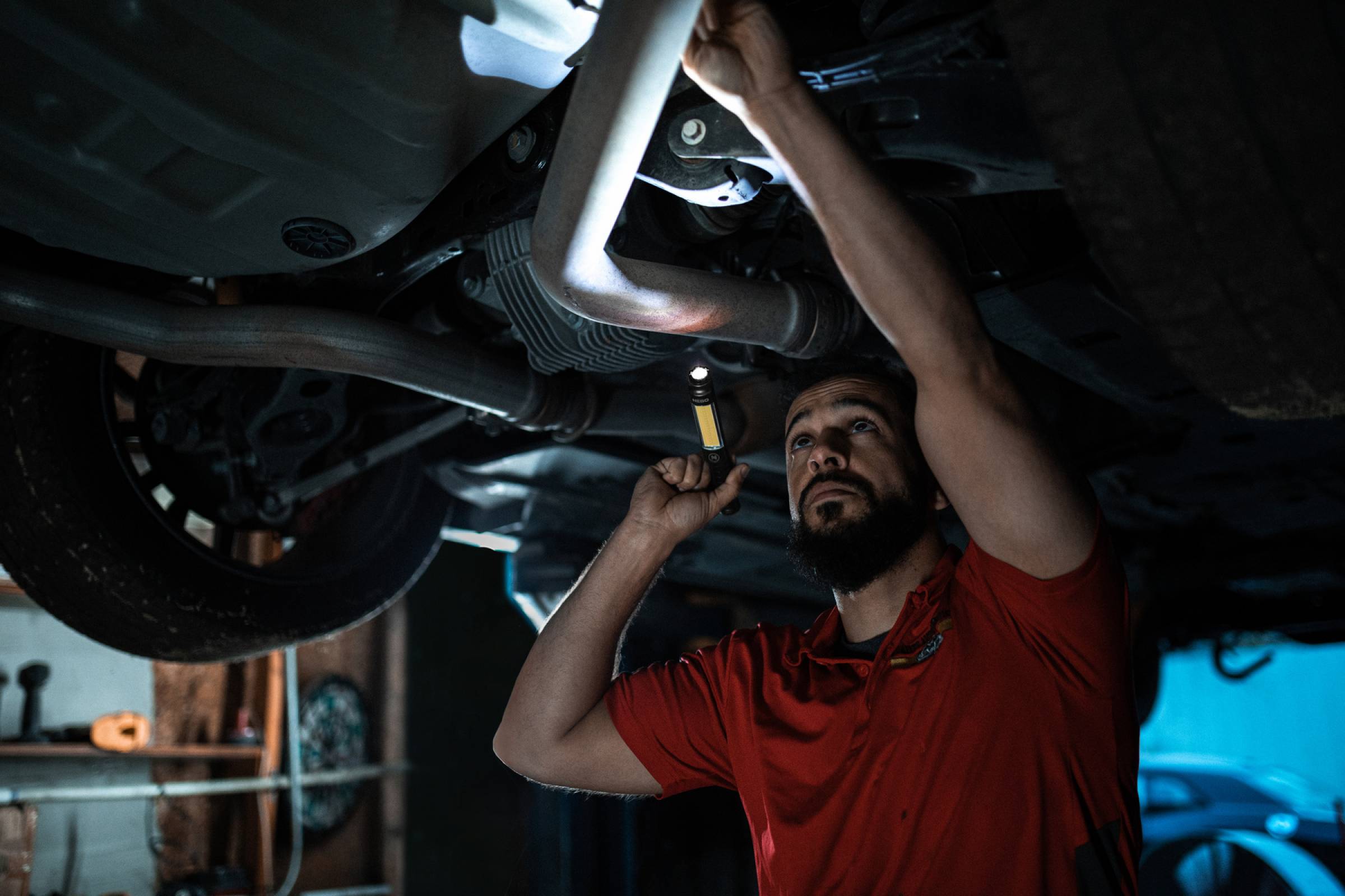 Ein Mechaniker in einem roten Hemd arbeitet unter einem angehobenen Auto und benutzt dabei eine Taschenlampe und ein Werkzeug in einer Werkstatt.