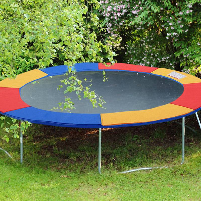 Ein rundes Trampolin mit einem mehrfarbigen Sicherheitspolster steht auf Gras, umgeben von grünem Laub, unter einem sonnigen Himmel.