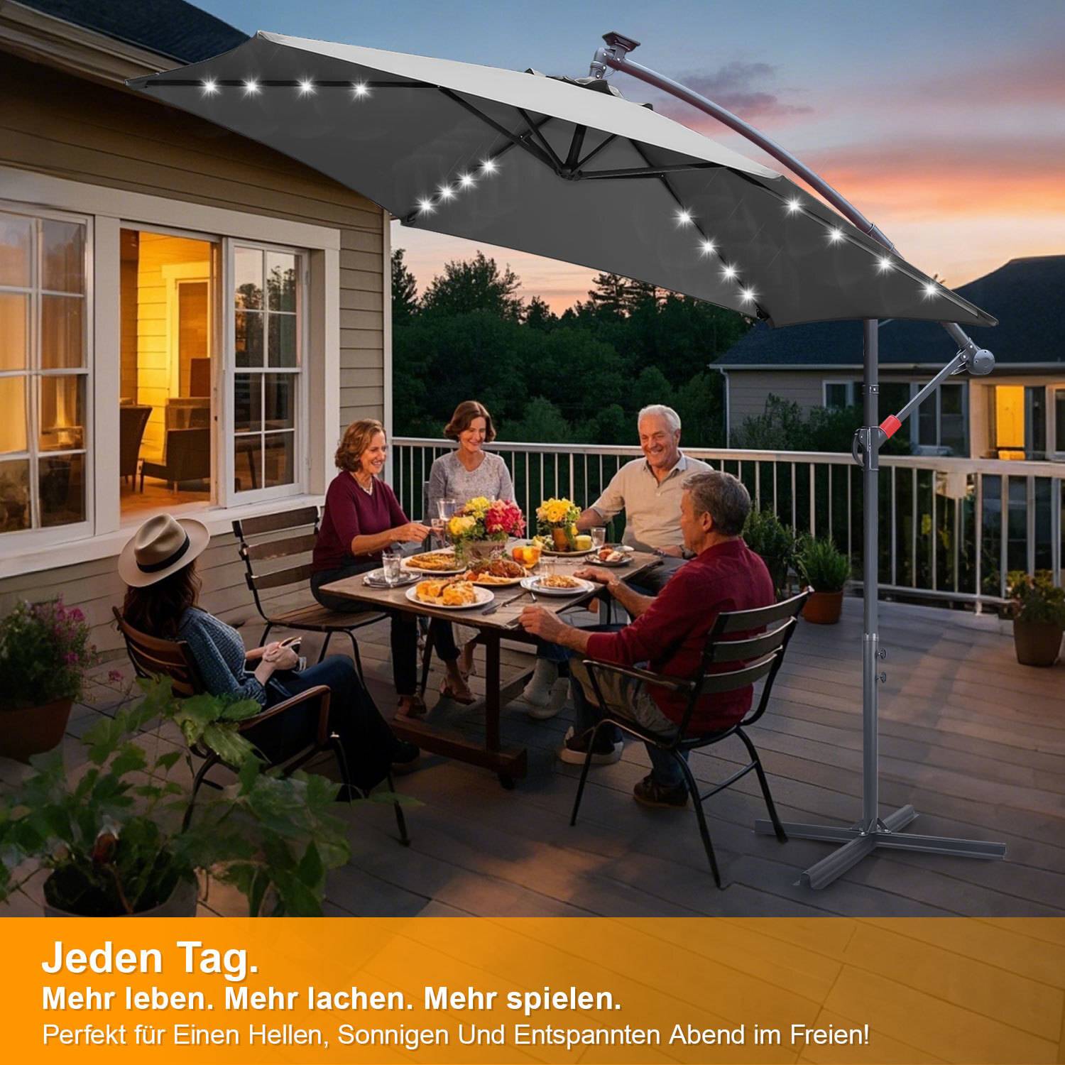 Fünf Personen sitzen an einem Esstisch auf einer Terrasse bei Sonnenuntergang, unter einem großen Sonnenschirm mit Lichterketten, und genießen Speisen und Getränke.