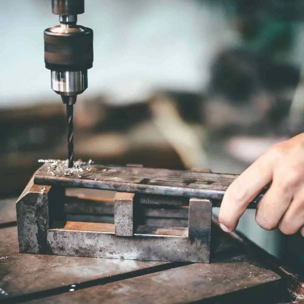 Eine Hand bedient eine Bohrmaschine, um ein Loch in ein in einem Schraubstock gehaltenes Metallstück zu bohren, wobei der Fokus auf Präzision in einer Werkstattumgebung liegt.