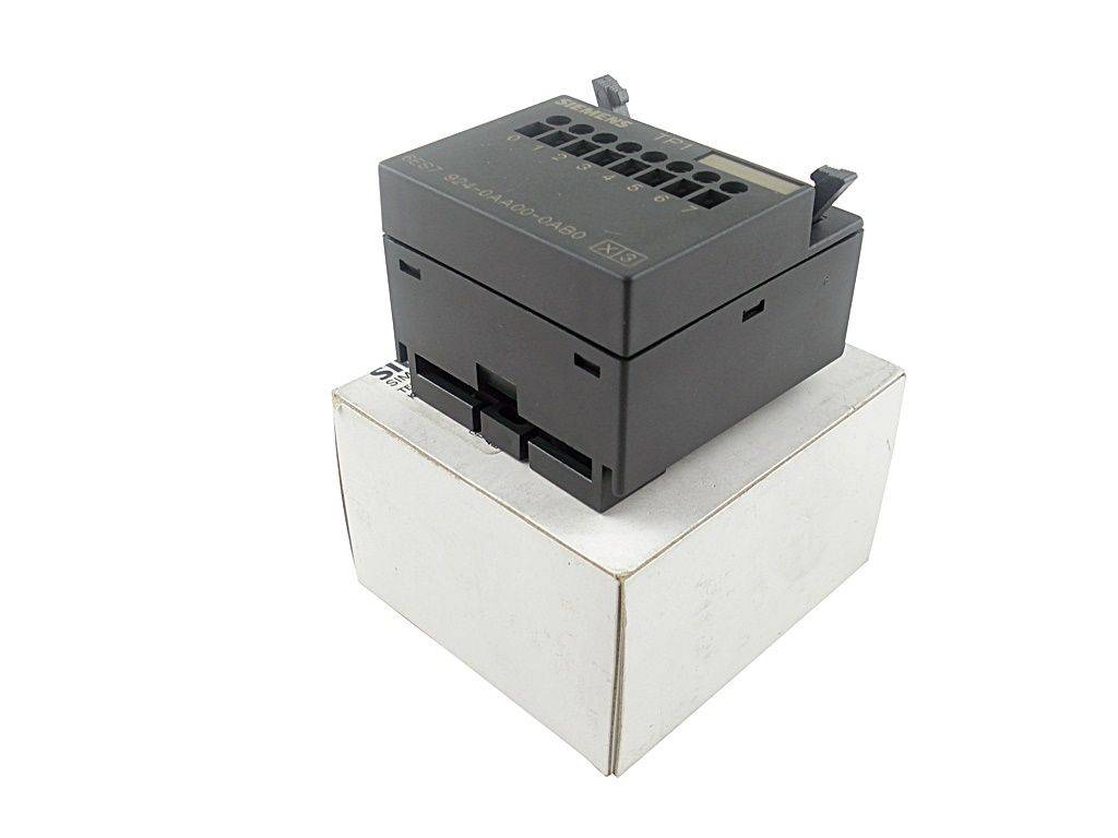 Eine schwarze elektronische Komponente sitzt auf einer weißen Box. Die Komponente verfügt über nummerierte Steckplätze und Anschlüsse, die wahrscheinlich in elektrischen Aufbauten verwendet werden.