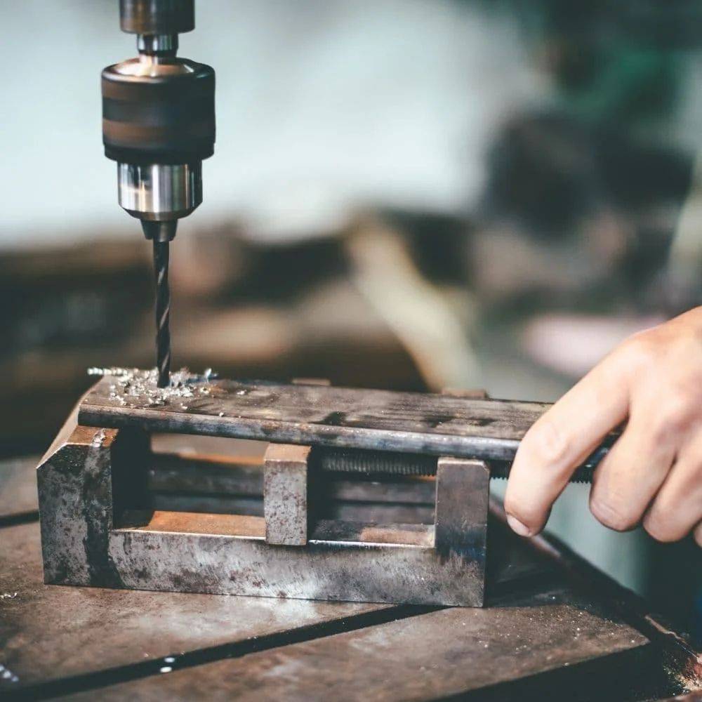Eine Hand bedient eine Bohrmaschine und bohrt ein Loch in ein Metallstück, das in einem Schraubstock befestigt ist, und demonstriert dabei einen Metallbearbeitungsprozess in Aktion.