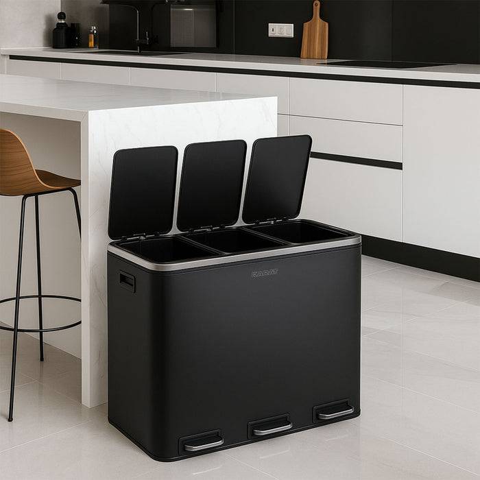 Eine moderne Küche mit einem schlanken schwarzen Mehrkammer-Mülleimer im Vordergrund, offen, um separate Recycling-Abschnitte zu zeigen. Arbeitsplatte und Hocker sind sichtbar.