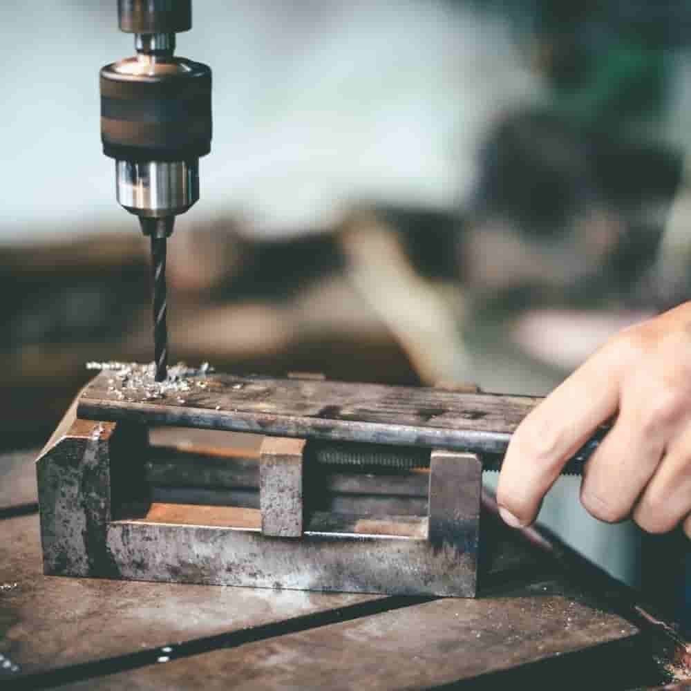 Die Hand einer Person bedient eine Metallbohrmaschine und bohrt in ein Metallstück, das sicher in einem Schraubstock eingespannt ist, was eine Werkstattaktivität demonstriert.