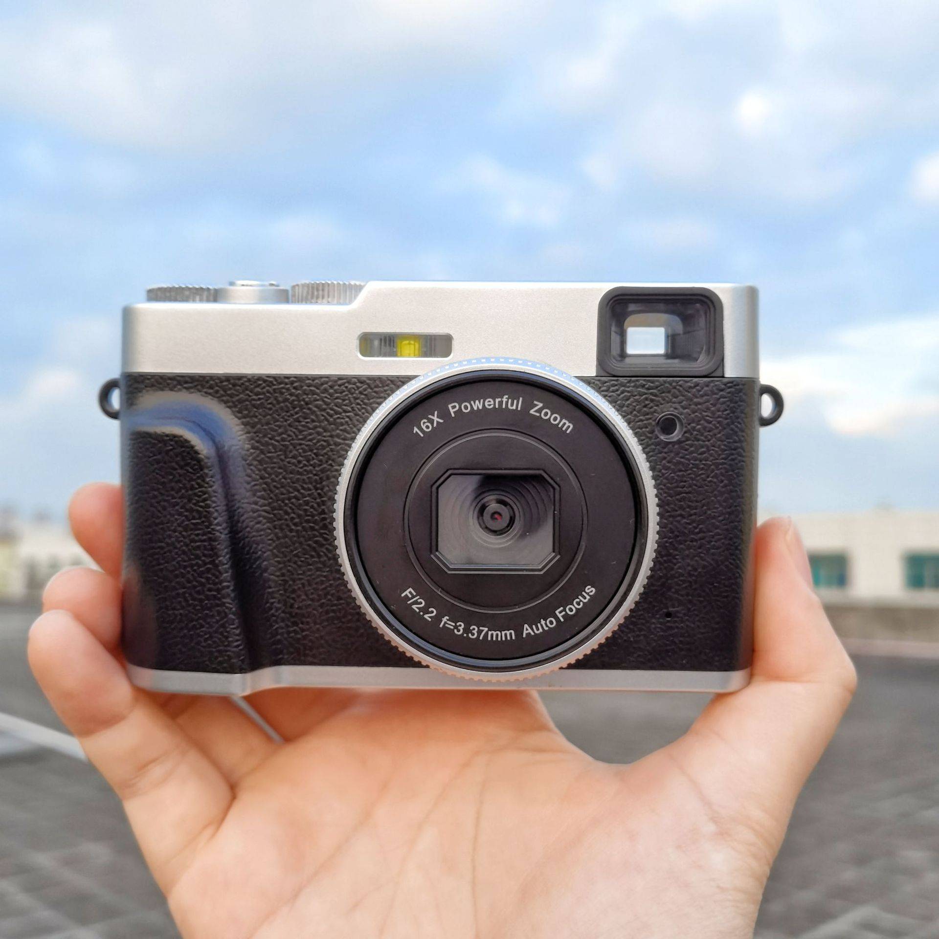 Eine Person hält eine Digitalkamera mit einem 16-fach starken Zoomobjektiv vor einem unscharfen Hintergrund aus bewölktem Himmel und Gebäudedächern.