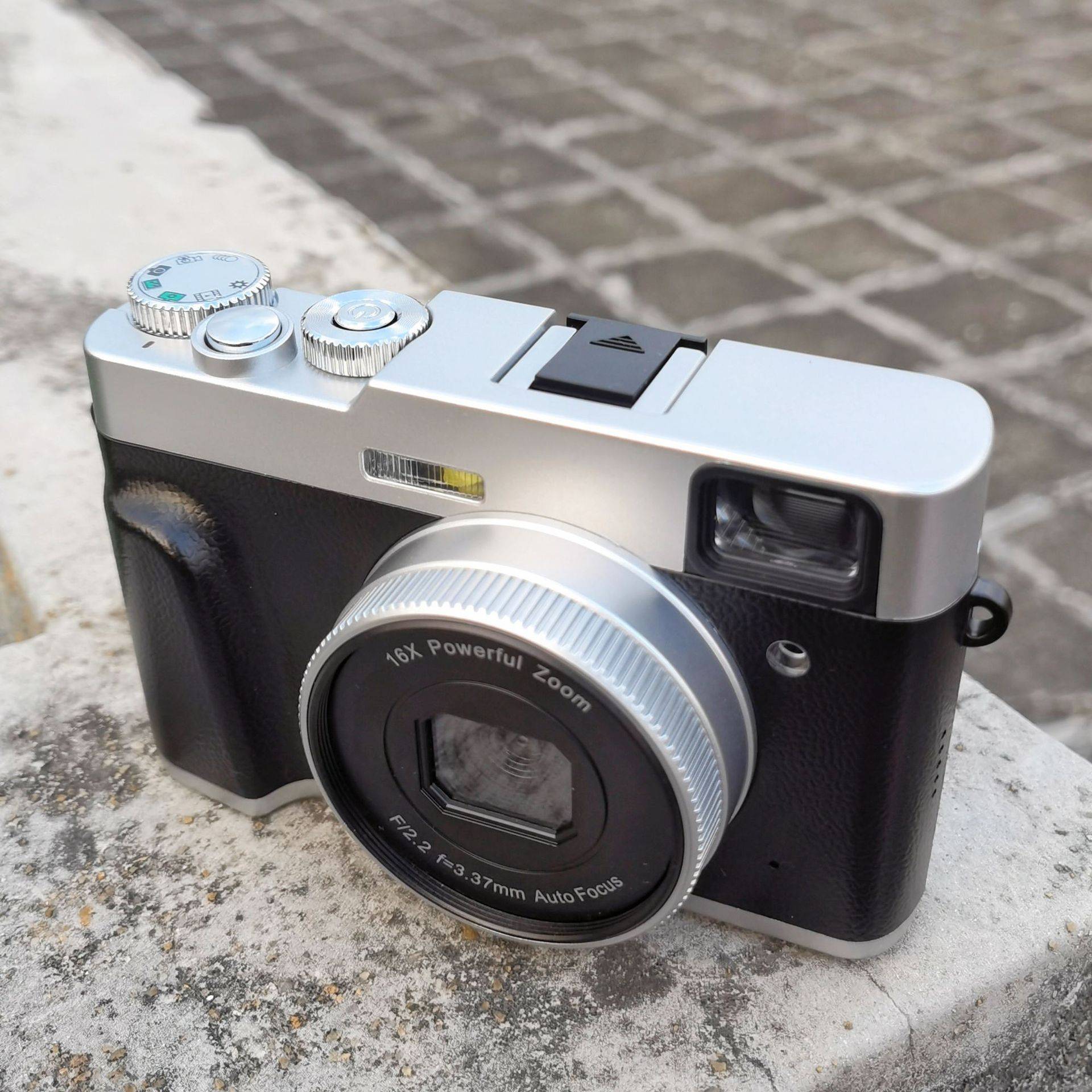 Eine Vintage-Stil-Kompaktkamera mit silbernem Gehäuse und schwarzem Griff sitzt auf einer Steinoberfläche und verfügt über ein Objektiv mit der Bezeichnung „16X Powerful Zoom