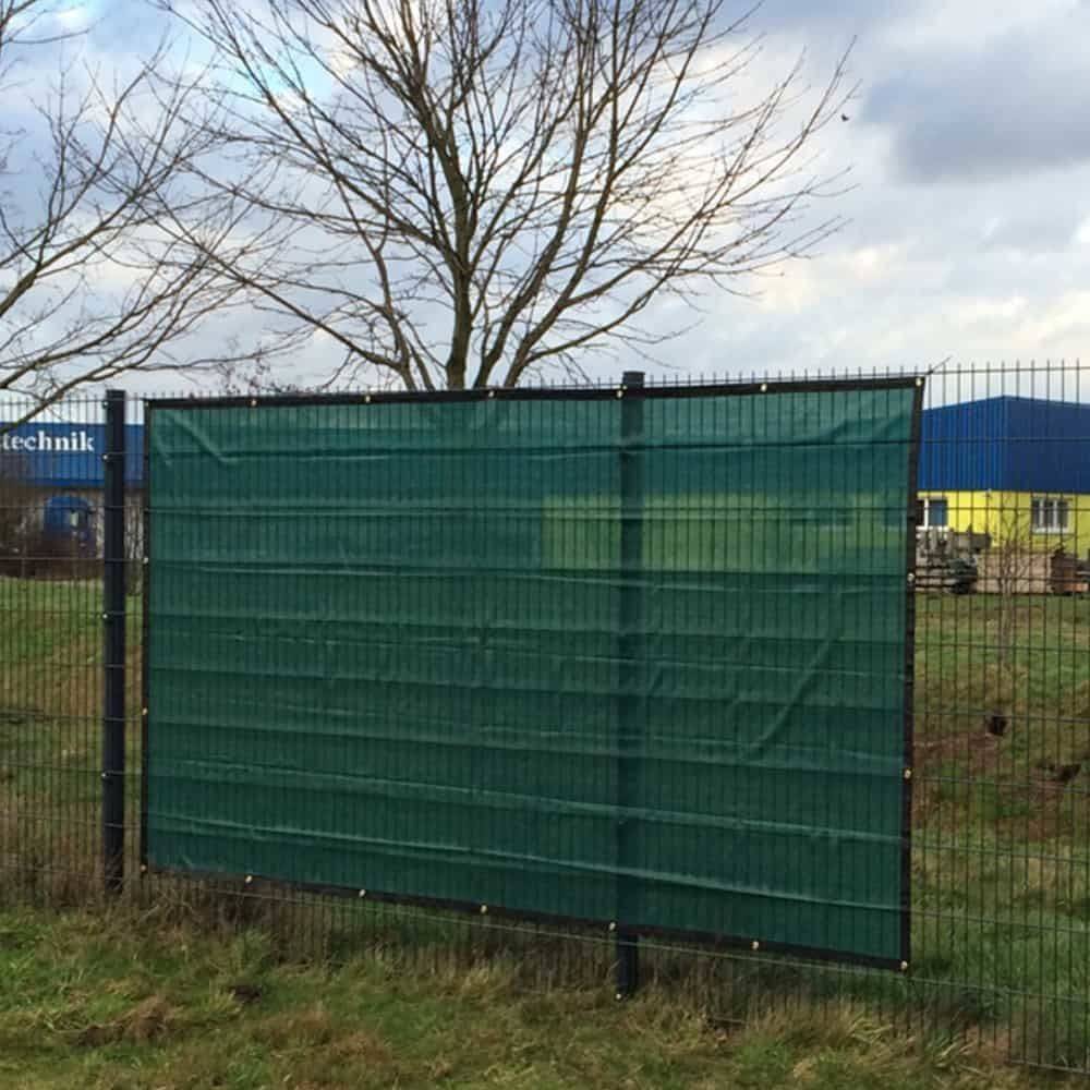 Ein Metallzaun mit einer grünen Stoffbespannung steht in einem Grasbereich, mit kahlen Bäumen und Industriegebäuden im Hintergrund.