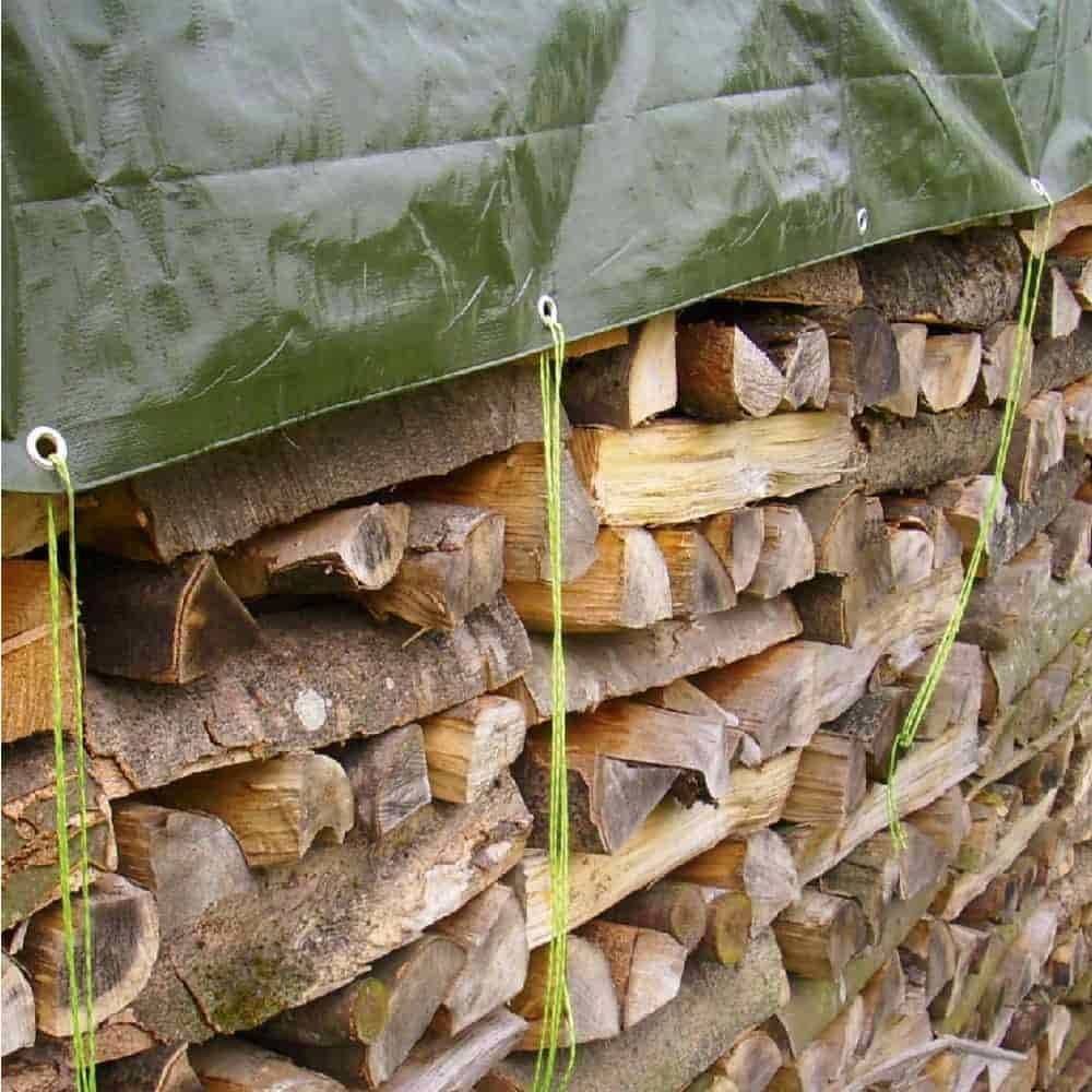 Ein Stapel gehackten Brennholzes ist ordentlich gestapelt und mit einer grünen Plane bedeckt, die durch gebundene Seile gesichert ist und einen Schutz vor Witterungseinflüssen andeutet.