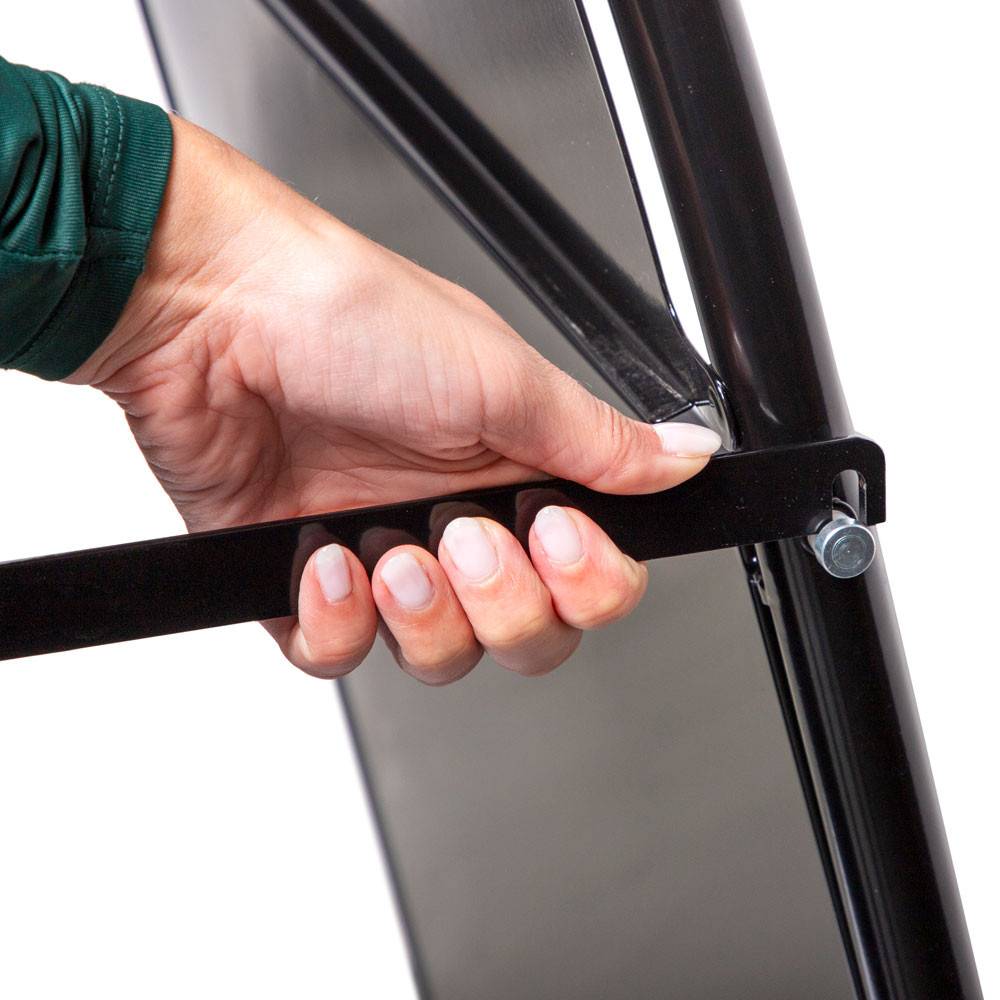 Eine Person mit grünem Ärmel betätigt einen Klappmechanismus an einer schwarzen Metallstaffelei und zeigt, wie man den Ständer zusammenklappen oder verstellen kann.