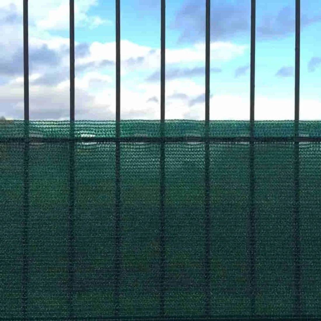 Ein hohes grünes Maschendrahtzaun mit Metallstangen oben vor einem bewölkten blauen Himmel.